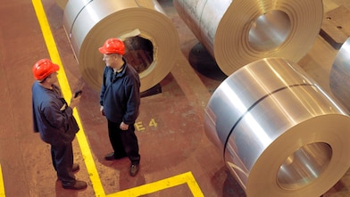 Two factory workers talking next to large rolls of steel