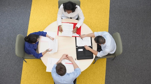 Four business people sitting around a round table reading documents, grey and yellow carpet