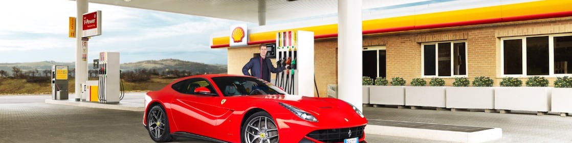 A red Ferrari sitting on a Shell station forecourt with a man leaning on a petrol pump